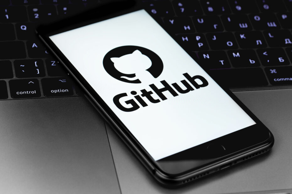 A mobile phone is sitting on a computer keyboard. On the screen is an image of the GitHub logo.